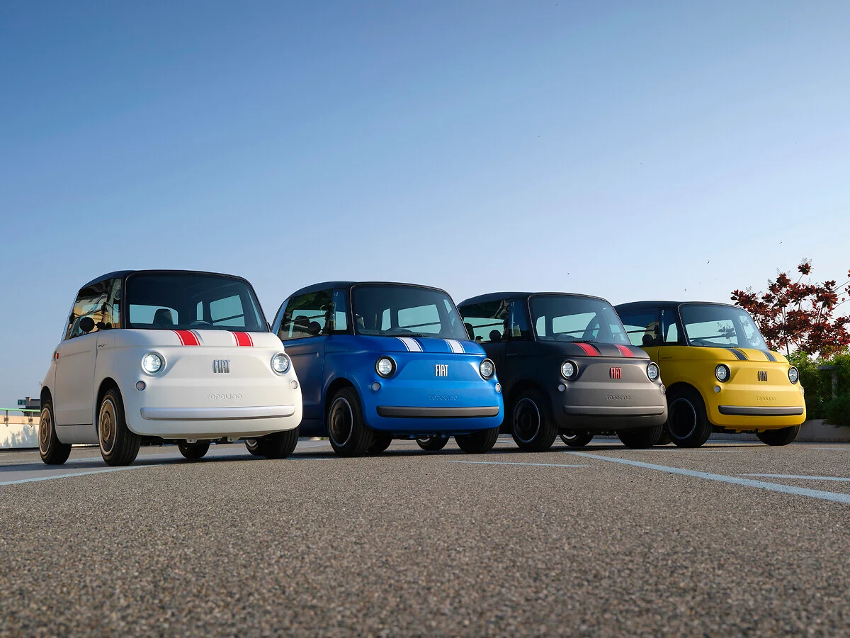 Vier compacte Fiat Topolino's Sport naast elkaar opgesteld op een parkeerterrein. Van links naar rechts zijn ze uitgevoerd in wit met rode strepen, blauw met witte strepen, donkergrijs met rode strepen en geel met zwarte strepen. Op de achtergrond een heldere blauwe lucht en rustige uitstraling.