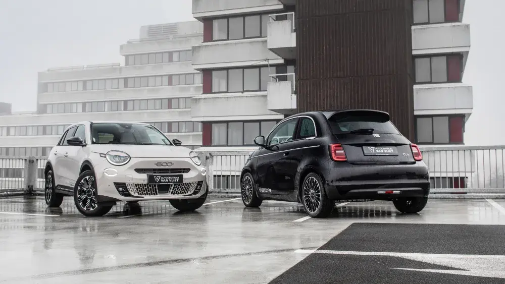 Een witte Fiat 600 en een zwarte Fiat 500 op een nat parkeerdek met mistige flats op de achtergrond.