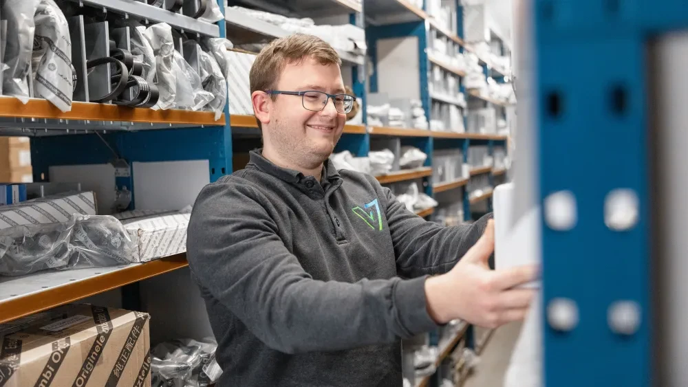 Een medewerker van Autocentrum Van Vliet glimlacht terwijl hij een auto-onderdeel vasthoudt in een geordend magazijn.