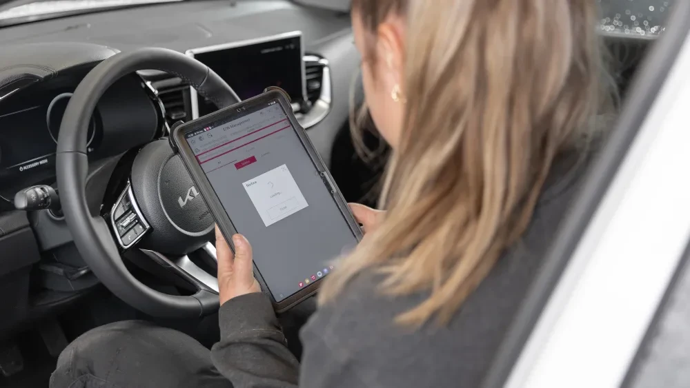 Een vrouw in een Kia voert een taak uit op een tablet, waarop een laadscherm zichtbaar is, ze lijkt bezig met voertuiginspectie of -onderhoud.
