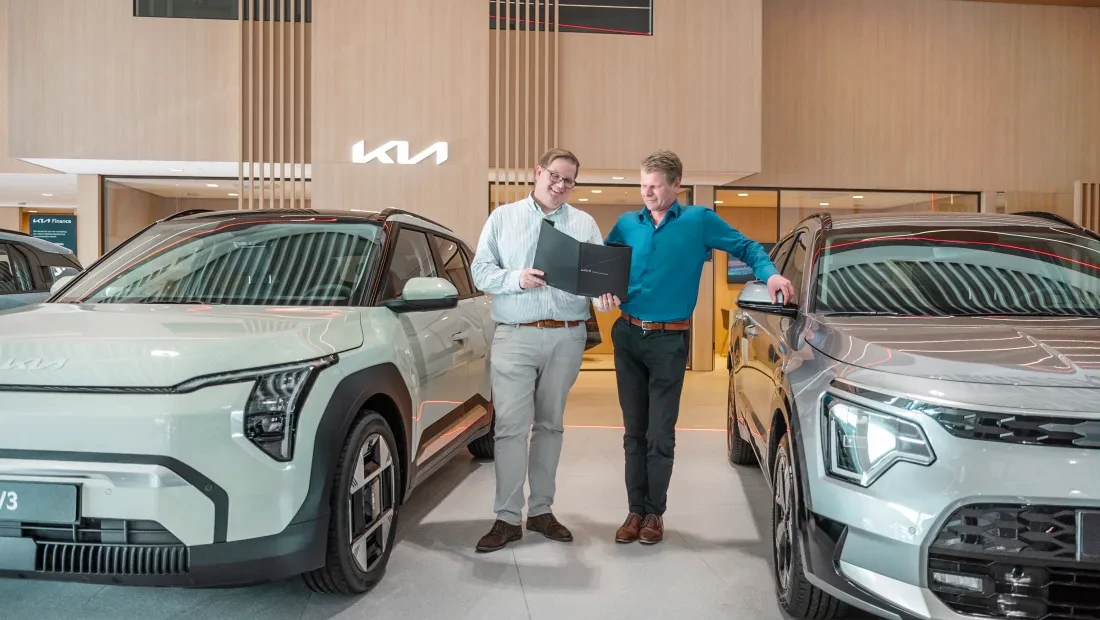 Twee verkoopadviseurs bij een Kia-dealer bespreken een auto-aankoop in een moderne showroom.